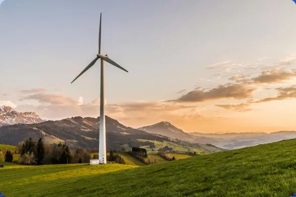 Wind turbine on a grassy hill at sunset with mountains in the background.